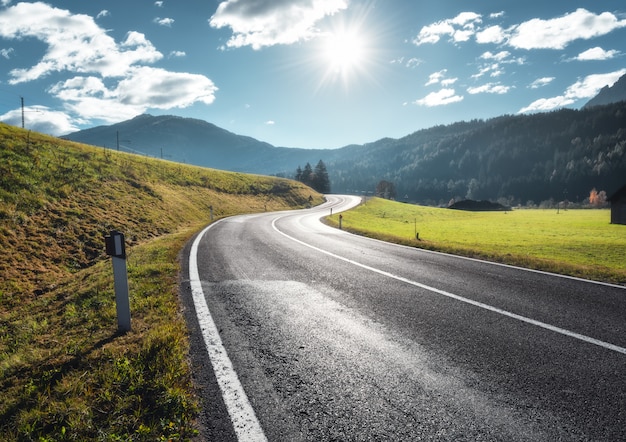 Road in mountain valley at sunny morning in Dolomites, Italy. View with asphalt roadway, meadows with green grass, mountains, blue sky with clouds and sun. Highway in fields. Trip in europe. Travel