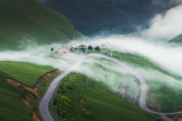 Road in the mountains in foggy evening. Clouds over the houses and green hills. Summer nature