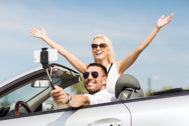 road trip, leisure, couple, technology and people concept - happy man and woman driving in cabriolet car and taking picture with smartphone on selfie stick