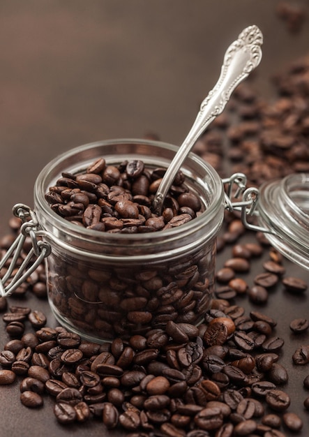 Roasted coffee beans in glass jar with steel spoon on brown background Macro