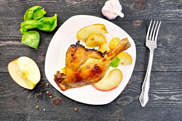 Roasted duck leg with apple, potatoes in a white plate, basil, garlic and fork on the background of the wooden planks on top