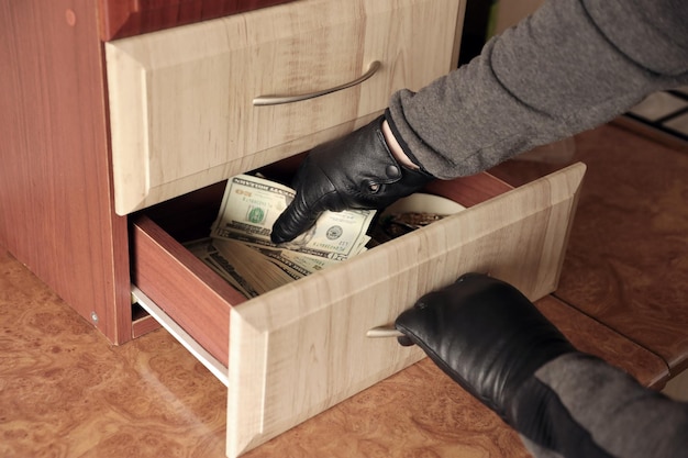 Robber in black outfit and gloves see in opened shelf in kitchen The thief takes out the US dollar bills from a shelf