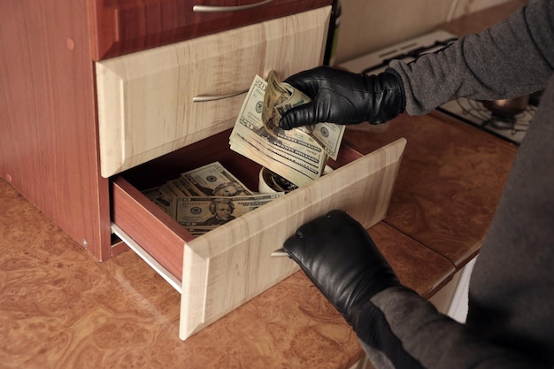 Robber in black outfit and gloves see in opened shelf in kitchen The thief takes out the US dollar bills from a shelf