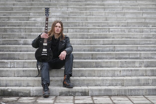 Rock guitarist on the steps. A musician with a bass guitar in a leather suit. Metalist with a guitar on the background of industrial step.