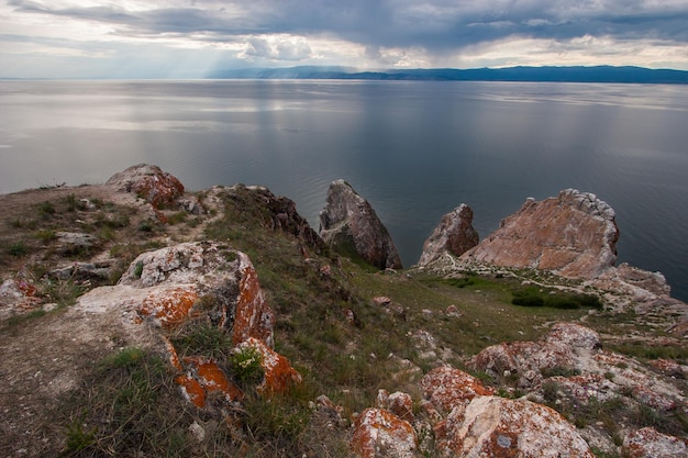 Rocks Three Brothers on the island of Olkhon on lake Baikal Sky with clouds On the stones is red moss and green grass around It is raining in the distance mountains are visible