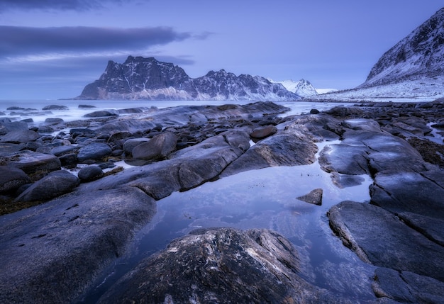 Rocky beach in winter Seashore with stones blurred water snowy mountains and violet sky with clouds at dusk Uttakleiv beach in Lofoten islands Norway Landscape with sea rocks at night Nature