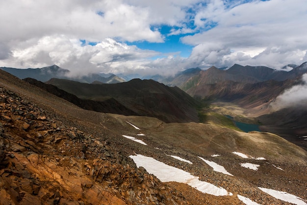 The rocky edge of a mountain pass and a view of a valley with a lake Blue sky and clouds Shadows from the clouds on the mountains Horizontal