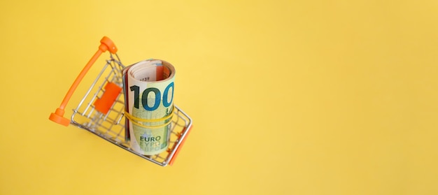 A roll of euro banknotes with a yellow elastic band in a small trolley on a yellow background.