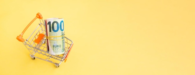 A roll of euro banknotes with a yellow elastic band in a small trolley on a yellow background.