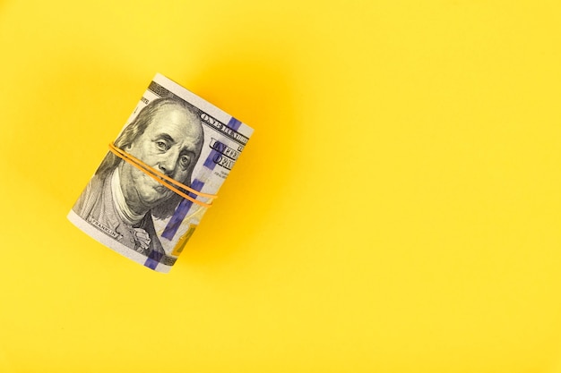 A roll of hundred dollar American bills is tied with an elastic band on a yellow background