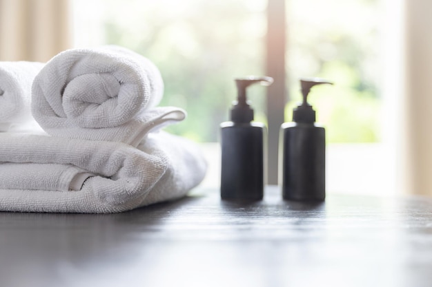 Roll up of white towels with shampoo bottles and liquid soap bottles on table with copy space on blurred room background For product display montage