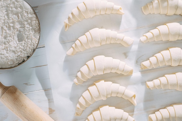 Rolled croissant dough and ready for baking on a white wooden table on baking paper in the morning sun