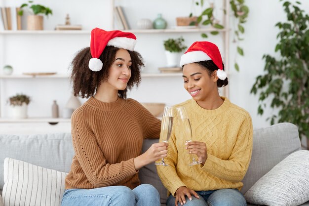 Romance date, celebrating Christmas and New Year with family or friends during covid-19 pandemic. Smiling african american young females in Santa hats clink glasses with champagne in cozy living room