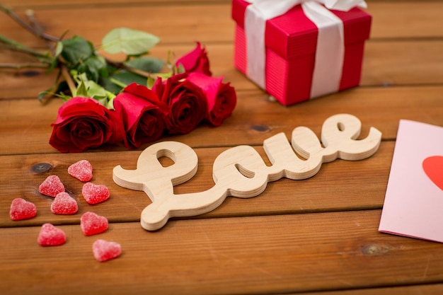 romance, valentines day and holidays concept - close up of word love, gift box, red roses and greeting card with heart-shaped candies on wood