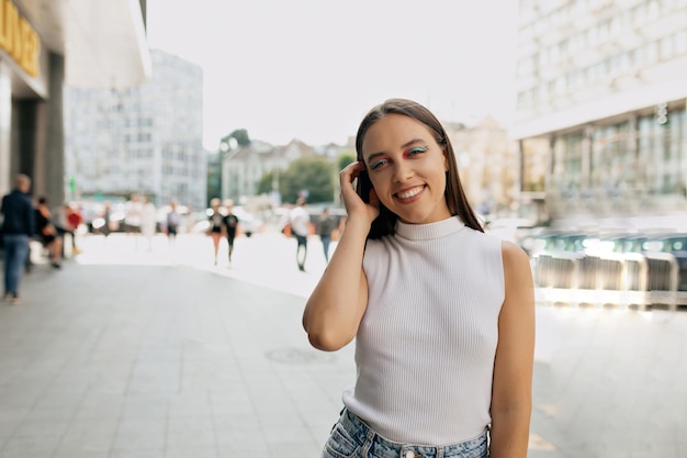 Romantic adorable girl with bright make up is touching hair and looking at camera with happy emotions dressed white tshirt and jeans