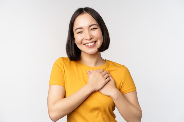 Romantic asian girfriend holding hands on heart smiling with care and tenderness standing in yellow tshirt over white background