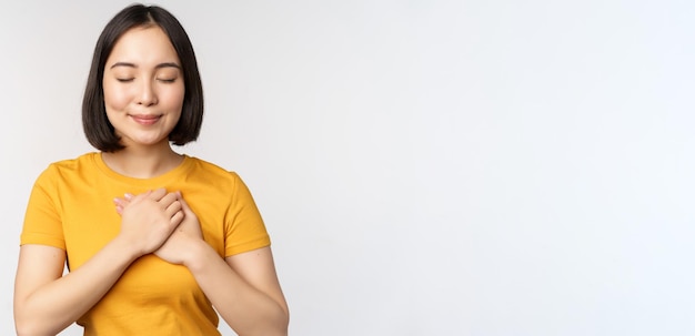 Romantic asian girfriend holding hands on heart smiling with care and tenderness standing in yellow ...