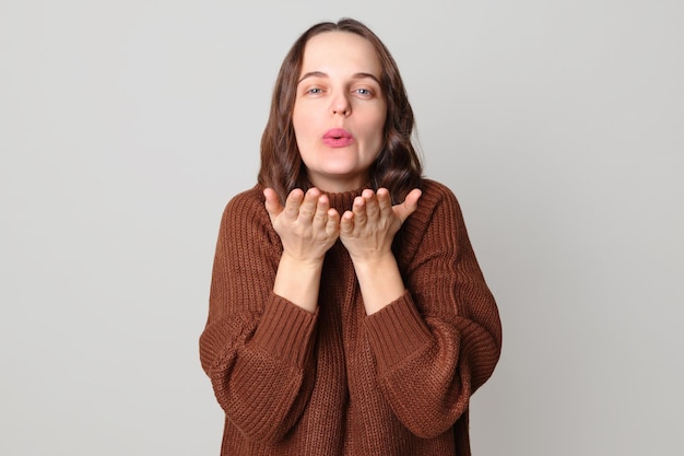 Photo romantic beautiful cute brown haired woman wearing brown jumper standing isolated over light gray background flirting with her boyfriend sending air kisses over palms