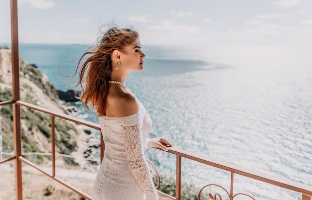 Romantic bride blond girl in white wedding dress with open shoulders posing on open terrace with backdrop of the sea and rocks Stylish young woman standing on terrace and looking on ocean at sunset