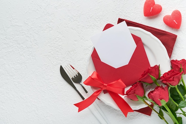 Romantic creative table setting Empty white plate and red roses candles knife fork and decorative silk hearts on white background Valentines Wedding Birthday Womens and Mothers Day Flat lay