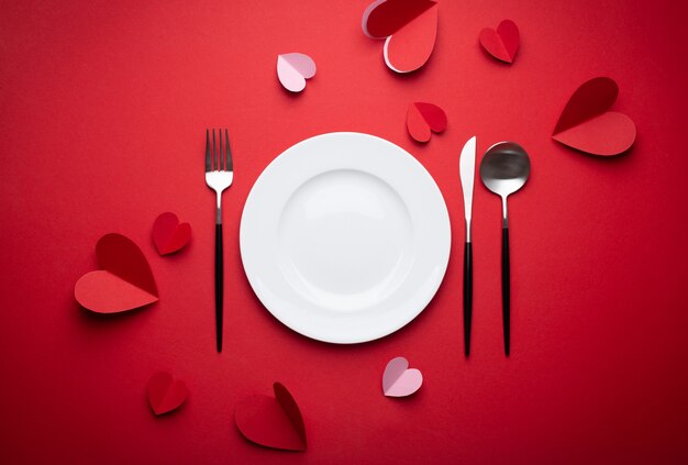 Romantic dinner at restaurant on Valentine's day, paper art. White empty plate, spoon, fork, knife on red background with paper hearts. Valentine's day date concept craft origami, space for text