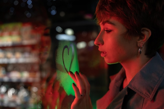 Romantic European woman in autumn coat draws heart on glass of evening cafe to confess love