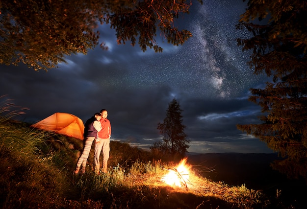 Romantic evening of a young couple hikers in the mountains by the fire under the starry sky in night at camping. Young people tourists standing near illuminated orange tent and looking at the fire.