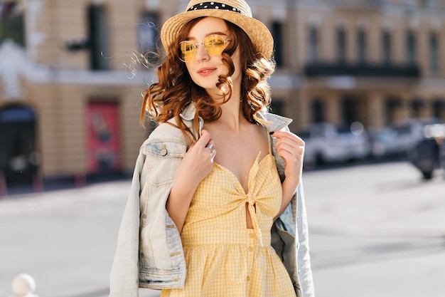 Romantic ginger girl in sunglasses walking around town Outdoor photo of spectacular european lady in summer hat and yellow dress