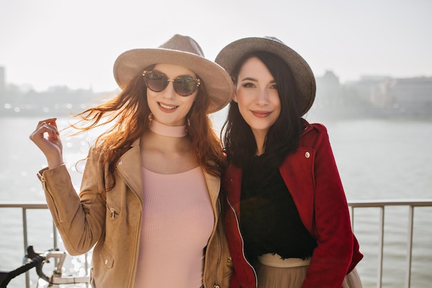 Romantic lady in beige jacket plays with long hair while posing with friend. Two white girls in hats enjoying sea views.