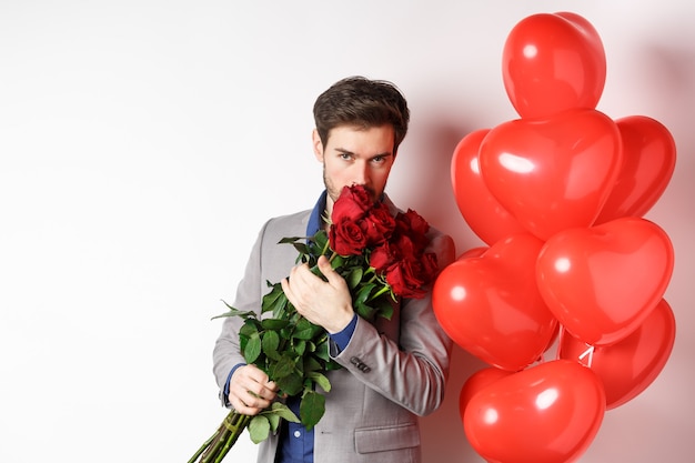 Romantic man smell bouquet of red roses and looking passionate at camera. Boyfriend in suit going on Valentines date with gifts and heart balloons, white background.