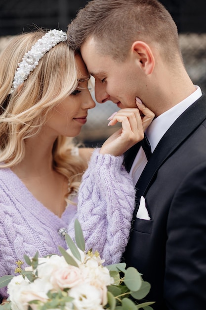 Romantic photo of young woman with blonde curly hair in lilac jacket, having bandeau on head and bouquet and man in black jacket and white shirt standing close with their foreheads touching outdoors