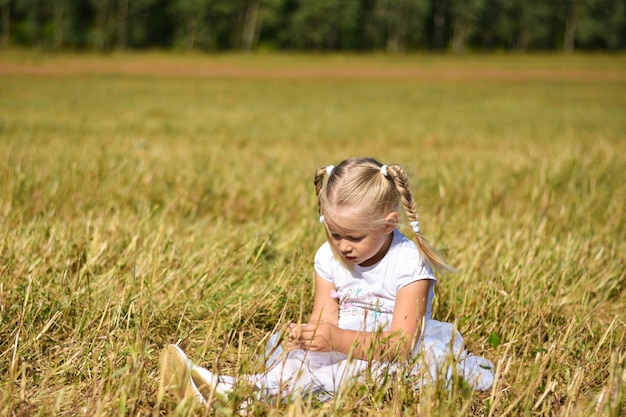 Romantic sad little girl in white dress sits on the grass in the field, looks down at hands