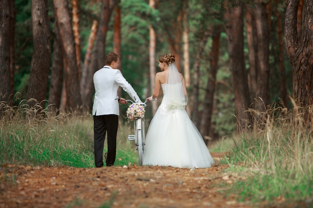 Romantic walk of bride and groom outdoors with bicycle. happy wedding couple in love walking and looking each other in the nature with bike. wedding day  in forest in summer. wedding