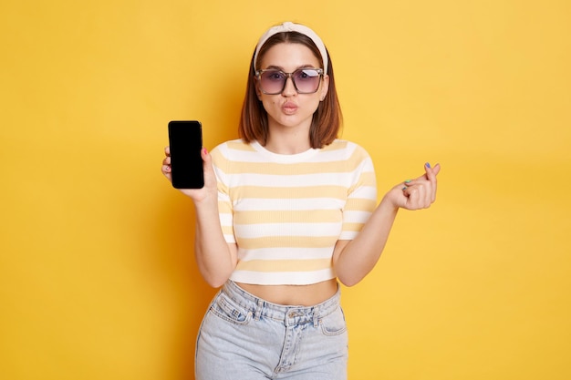 Romantic woman wearing striped shirt posing isolated over yellow background showing smart phone with advertisement area on blank scree sending air kisses making love korean gesture