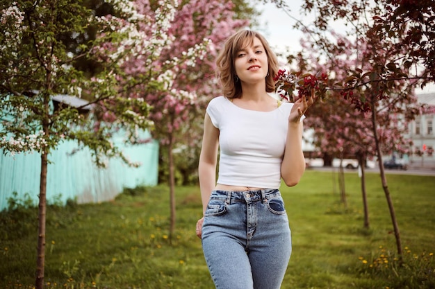 Romantic woman in white crop top and blue jeans outdoors in spring among blooming trees