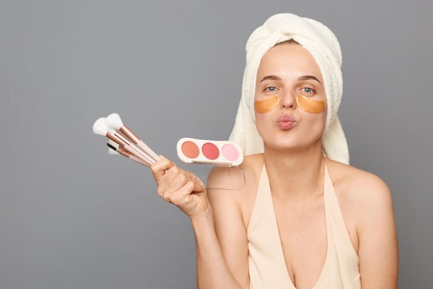 Romantic woman with patches under eyes wrapped in towel holding powder and brush isolated over gray background sending air kissing copy space for promotional text