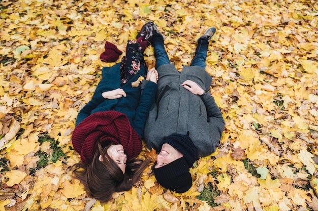 Romantic young couple lie on yellow leaves in park hold hands together look at each other with great love enjoy autumn sunny day People relationships togetherness and romance concept