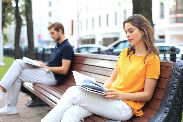 Romantic young couple in summer clothes smiling and reading books together while sitting on bench in city street.