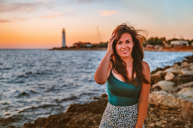 A romantic young woman in a skirt admires the sunset on the sea with a background lighthouse