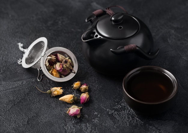 Rose buds mix tea in strainer infuser and ceramic teapot and cup on dark background