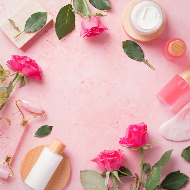 Rose extract natural cosmetics in reusable bottles with gua sha facial beauty roller and natural rose flowers on pink square frame