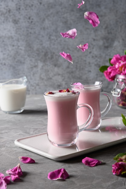 Rose moon milk in glass cups and falling rose petals on grey. Top view.