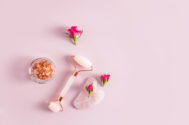 Rose quartz massager gua sha scraper salt in a bowl and small rosebuds Top view Pink background A copy space natural care