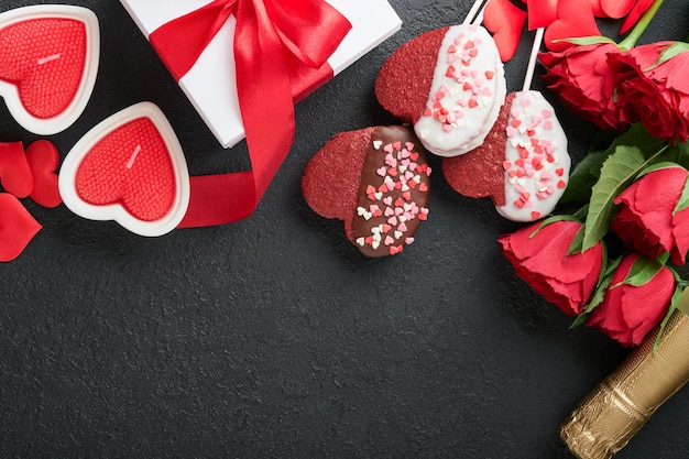 Rose red flowers brownie cookies and chocolate candy on heart shaped gift box with wine on black background Valentines day Mothers or Womens Day greeting card Top view flat lay with copy space