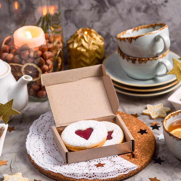 Round biscuits with heart-shaped jam in a gift box among Christmas decorations and candles on a gray table