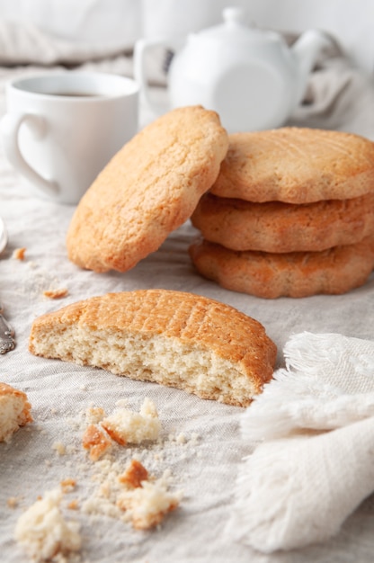 Round homemade shortbread cookies. Broken in half. On a linen tablecloth of gray color. In the background is a stack of cookies. White teapot and mug.