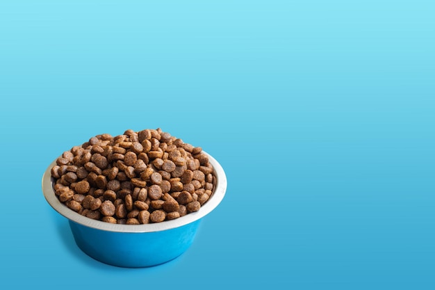 Round metal bowl full of crunchy dry food for dogs or cats on blue background, side view, copy space. Pet care concept. Delicious and healthy food for domestic cats and dogs. Diet food for pets.