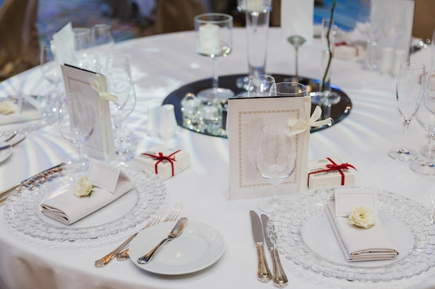 Round table served with empty plates knives forks card and napkins covered with white tableclothes Cutlery glassware on festive table