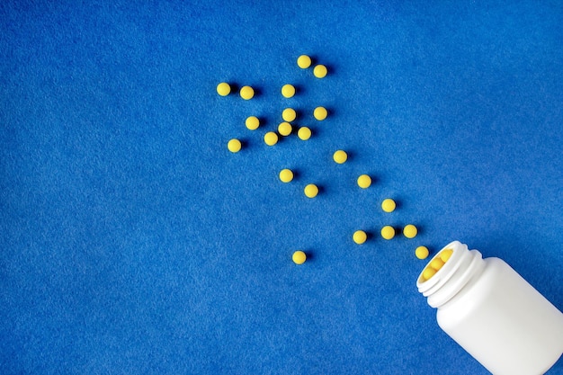 Round yellow vitamins spilled out of a white jar on a blue background copy space