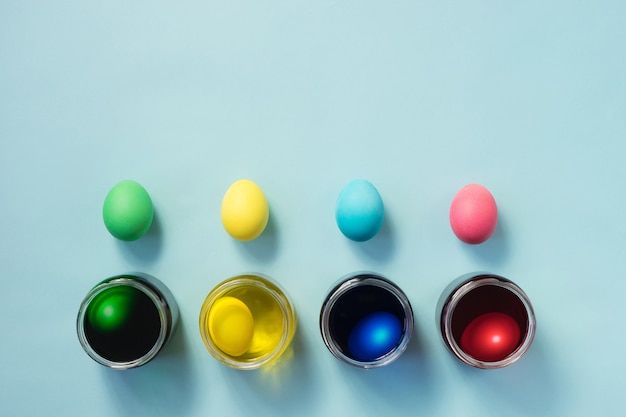 Row of colored easter eggs and a jars full of egg dye on pastel blue background. Flat lay.